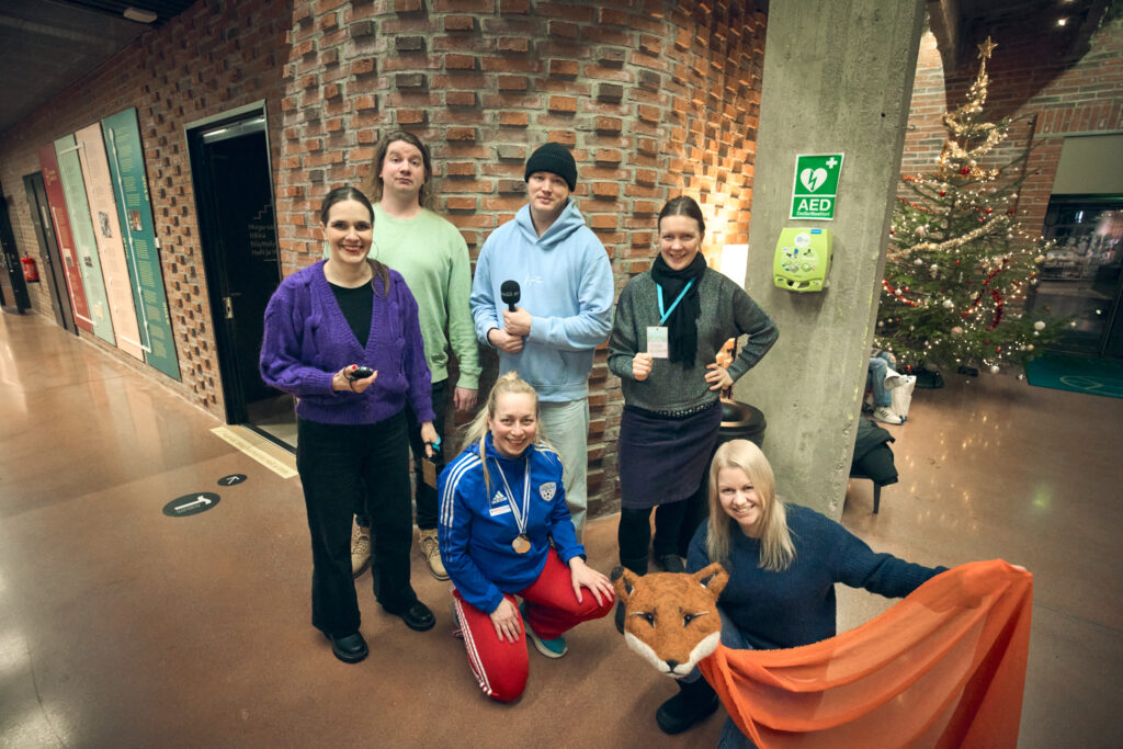 Kuusi ihmistä ryhmämuodostelmassa katsovat kameraan hymyillen Taide- ja kulttuurikeskus Kalevan Navetan autassa. Yhdellä kädessä mikrofoni, yhdellä kettu-nukke.